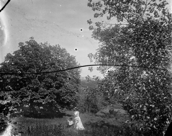 Syl and aunt Helen walking among trees on a hill. Behind them the roof and chimney of a house are visible.