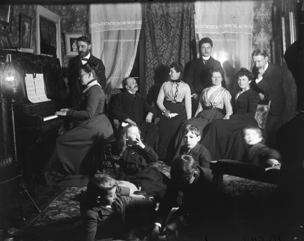 A large group of people are sitting around a woman playing a piano at night. Lamps light the scene, and children are playing on the floor.