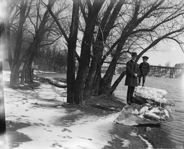 Syl and Will stand on ice covered rocks on a riverbank near some trees. Snow is on the ground and a house and bridge are in the background.