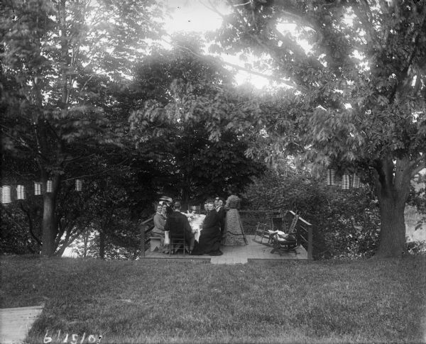 View from across lawn of Mrs. Woshbun with other women sitting around a table on a deck built on the edge of a hill overlooking a river. There are also rocking chairs on the deck and a string of paper lanterns in the trees.