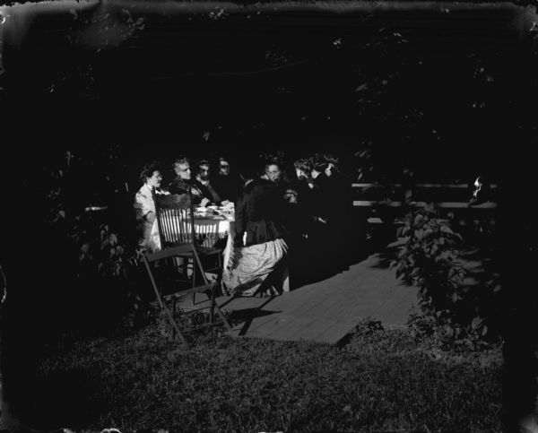 Mrs. Woshbun with other women sitting around a table on a deck built on the edge of a hill.