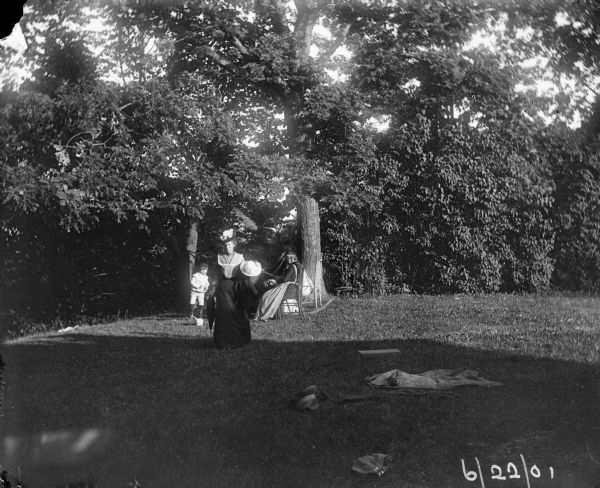 View across lawn of Syl and two other women near two trees strung with a hammock. One of the woman is walking away carrying a hat and umbrella. The lawn in the foreground is strewn with clothing.