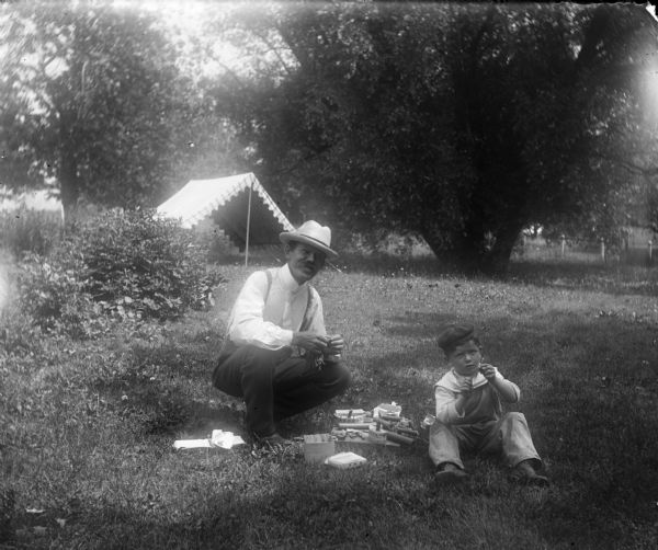 Syl and Pa, both wearing hats, sit on a lawn lighting fireworks. There is a tent or open-sided awning behind them among trees.