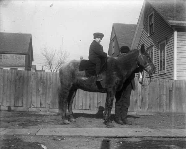 Side view of Syl sitting on the back of a small horse, while a handler stands behind him holding the reins. In the background is a wooden fence, with the tops of houses visible beyond it. In the foreground is a sidewalk.