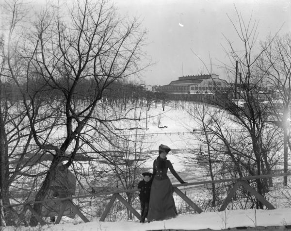 Syl and aunt Helen lean on a wooden hand rail on Gully bridge. Behind them is a lake with ice and snow. A large factory-like building is visible in the distance on the far shoreline.