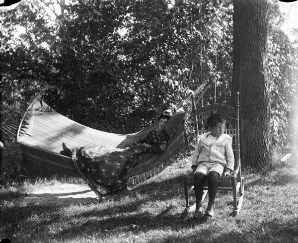 Aunt Helen lounging on a hammock slung between two trees. Her son, Syl, is sitting nearby on a cane rocking chair.