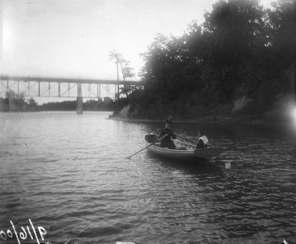 Syl and aunt Helen in a rowboat on the Milwaukee River at Gordon Park. In the background a large bridge crosses the river. On the right the banks of the river are covered with foliage.