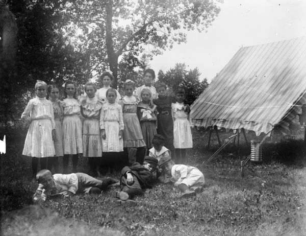 A group portrait of aunt Helen's Sunday school class outside. There is a tent or open-sided awning to the right of the group. Nine girls stand in a line, while four boys recline on the grass in front of them.