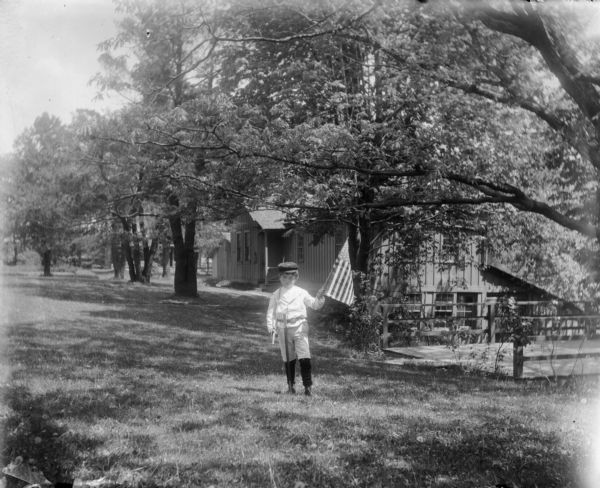 Syl standing on lawn with a sword in his right hand and an American flag raised in his left. Cabins are visible in the background on the right, as well as a deck suspended over a steep bank.
