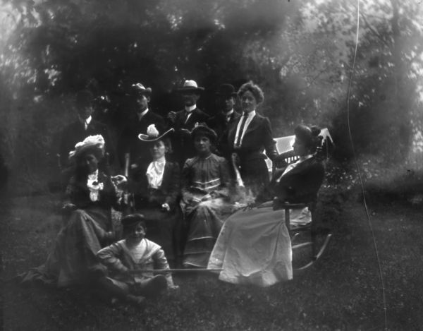 Group portrait of lawn party. Syl sits on the ground holding a sword in front of four women who are seated and one woman who is standing. The men, all wearing hats, are standing behind them.