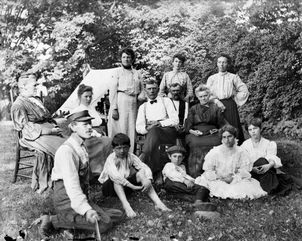 Family portrait of a large group of people posed on a lawn. In the background is a tent or open-sided awning. Harry Dankoler, wearing a hat a eyeglasses, is seated on the ground on the left, with Syl directly to the right of him. Harry is holding a string or rope in his right hand, which is a shutter release mechanism.