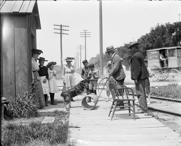 Syl and other children watch a dog perform with a barrel on a wooden sidewalk near a small building. Railroad tracks cross behind them, and power lines stretch to the horizon. A covered wagon is going by on a road on the right.