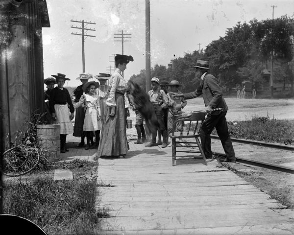 A group of children watching a performing dog jump towards aunt Helen. Railroad tracks, power lines and horse-drawn carriages are visible in he background.