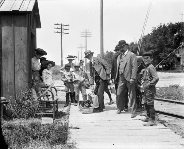 A group of people, Syl among them, watch a man with a performing dog in a wicker suitcase. Railroad tracks and power lines are in the background.