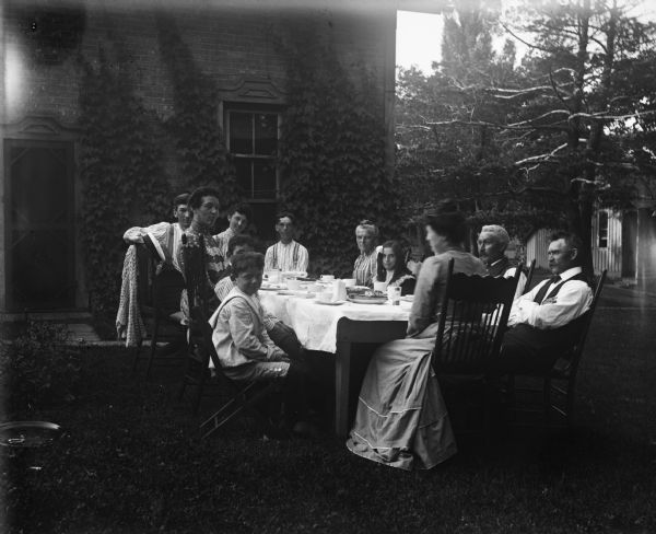 A group of children and adults sits around an outdoor table set with dishes just outside of a house covered in ivy. Syl is in the foreground grinning at the camera.