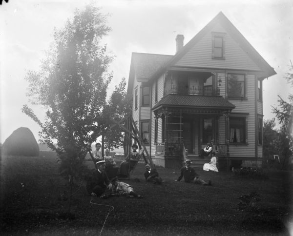 Group posed on the lawn of Uncle Herman's house. Three women are seated on a platform swing while others are sitting on the lawn and the porch. Harry Dankoler is seated on the ground on the far left with Syl at his side. Harry holds a shutter release mechanism in his right hand.