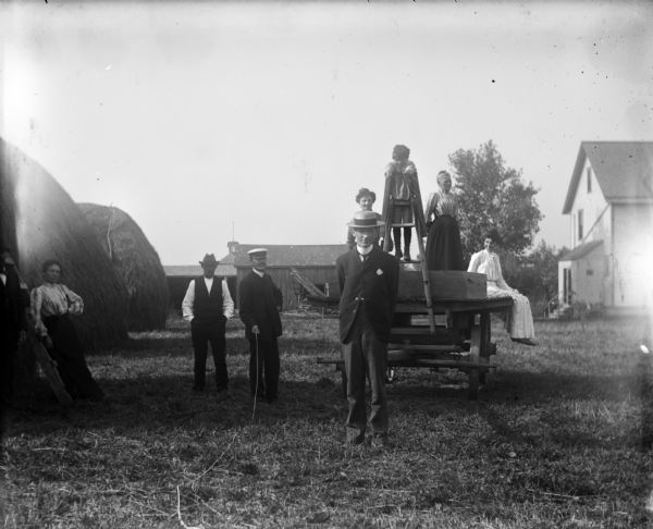 Group of well-dressed people posing outdoors at Uncle Herman's farm. Harry Dankoler is the man in the middle holding the shutter release mechanism. On the right are large haystacks, and in the background are farm buildings and a farmhouse.