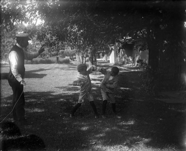 Harry Dankoler, wearing a hat and holding the shutter release mechanism in his right hand, is stepping in to stop a possibly posed fight between Syl and Barney outdoors on a lawn.