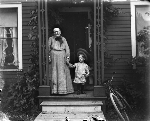 A very young Syl wearing a wide brimmed hat stands holding hands with his grandmother on a front stoop. A bicycle is leaning up against the porch to the right.