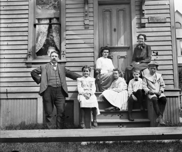 View over fence of a group of people and one dog seated on steps leading up to the door of a house. Syl is seated on the far right of the group wearing a hat. A sign to the right of the door says: "This Property for Sale."