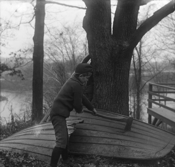 Syl chopping the hull of an overturned row boat which is leaning against a tree. Behind him is a steep cliff with a river or lake below, and on the right is a deck overhanging the cliff.