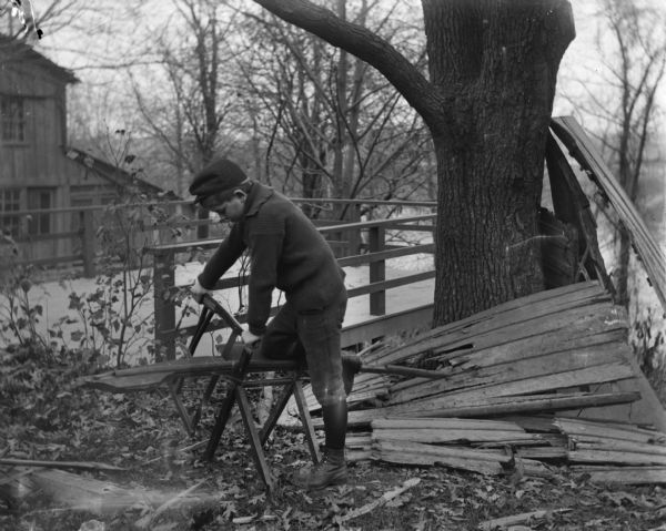 Syl sawing wood from the hull of an overturned rowboat on a small sawhorse. Behind him is a deck overhanging a steep cliff with a river or lake below. A house is on the left behind the deck.