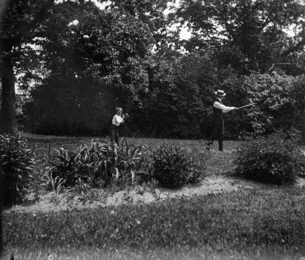 Syl and an unidentified man play baseball on the lawn. Syl is on the left wearing a catchers mask and glove, while the man is holding a bat preparing to hit a ball from an unseen pitcher.
