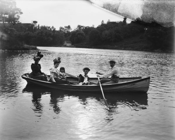 Mr. Hoose is rowing Syl, Ma, Mrs. Miller, Herbert, and Alice up river in a small rowboat. The women are wearing elaborate hats and there's a small boathouse visible in the background on the far shoreline. On the hill in the background is a tower near what may be a large industrial building.