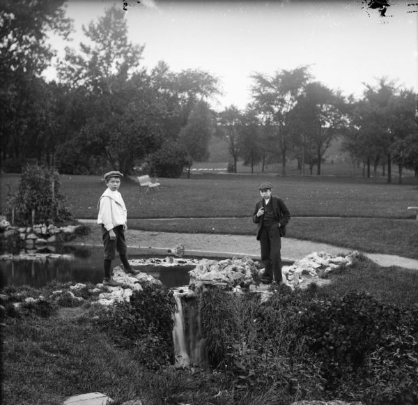 Syl and another boy stand near a small waterfall at Reservoir Park. A bench can be seen in the background.