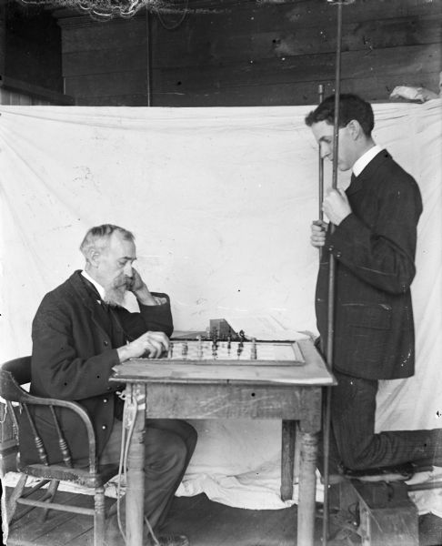 Two men pose playing a game of chess in front of a cloth backdrop in Dankoler's photography studio. The man on the right is standing and is holding what appears to be pool cues.