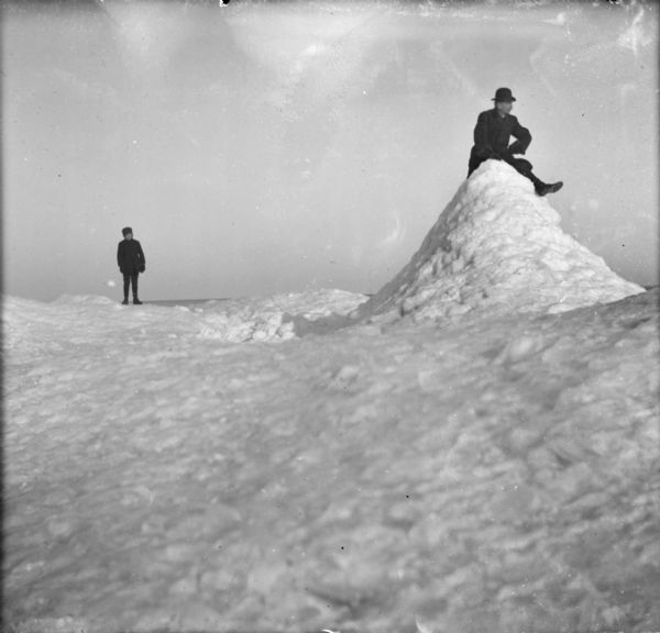 Winter scene with a man sitting on top of large pile of snow on the right, while Sylvester Dankoler (b. 1895) looks on from the background.
