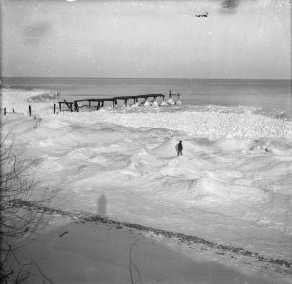 Elevated winter scene with Sylvester Dankoler (b. 1895) standing on ice and mounds of snow near a broken pier along a shoreline. The shadow of the photographer is visible on the lower left.