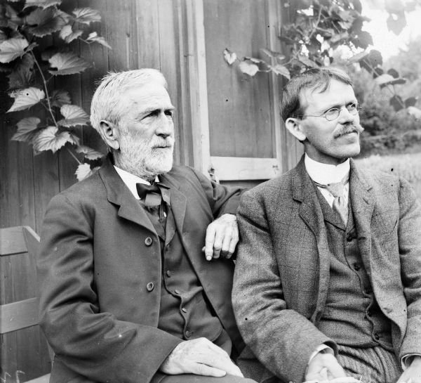 D.S. Crandall, on the left, is seated on a bench next to Harry Dankoler who is wearing eyeglasses. Behind them is the side of a house. Both men wear suit jackets, vests, and ties.