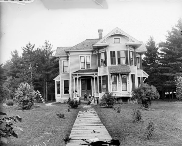 View down front walk of a small group of people with a dog seated on the porch of a frame house. There is a platform swing on the left in a side yard.