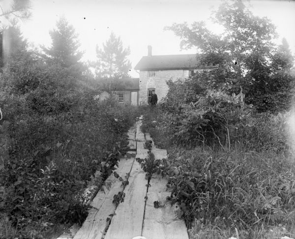 A path made of wooden planks leads through a tangle of shrubs and other small plants leading to Grandall's house, a two-story brick structure. A man stands in front of the house wearing a hat.