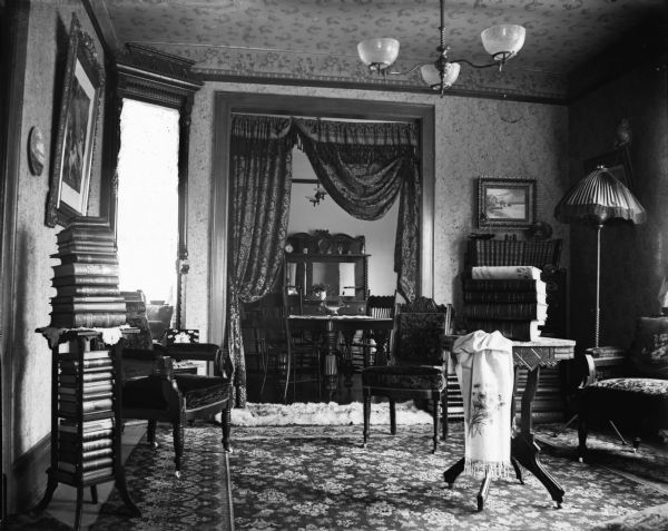 An interior view of a sitting room with a high ceiling, most likely Dankoler's. Books are stacked neatly on shelves and tables. Through an curtained archway the dining room is visible.