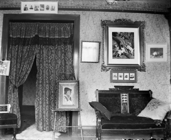 An interior view of a sitting room, most likely Dankoler's. There is a high-backed, upholstered, carved wood settee next to a framed picture resting on an easel in front of a curtained doorway.