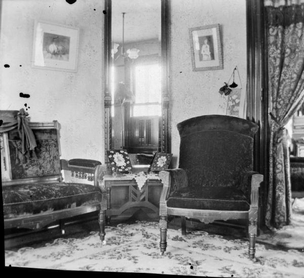 Interior view of the sitting room of the Dankoler's house. Framed pictures adorn the walls, and there is a tall mirror in the corner reflecting a window. A side table sits between a high-backed, upholstered, carved wood settee and a cushioned chair.