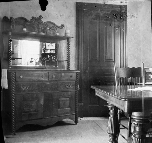 An interior view of the Dankoler's dining room. A carved cupboard with a mirror is on the left and on the right is a table and a chair.