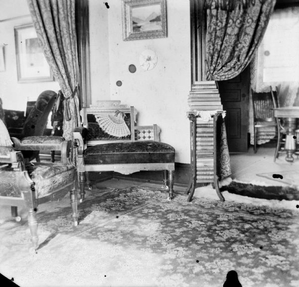 Interior view of the Dankoler's sitting room with cushioned chairs and books stacked on a side table. A deocorative hand fan rests on one of the chairs. Two curtained archways lead into rooms on the left and right.