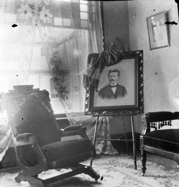 View of corner of the Dankoler's sitting room. A framed portrait of Harry Dankoler sits on an easel near a window with a lace curtain. On the left is a cushioned rocking chair.