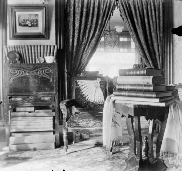 An interior view of the Dankoler's sitting room with books stacked under and on top of a carved wood cabinet and on top of a table. A cushioned chair sits in front of a curtained archway leading into another room.