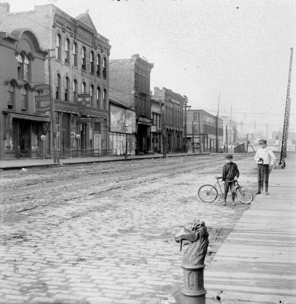 Syl stands holding an umbrella near an unidentified boy who is leaning on a bicycle on the edge of a cobblestone street. Storefronts are along the street on the left. In the foreground is a horse head hitching post. Signage on storefronts reads: "Lager Beer Hall F.Agenten" "Fred Miller Lager Beer" ans "Lodging." At the end of the street is a harbor with boats.