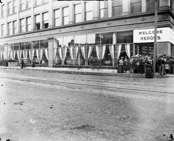 View from across street of the exterior of a large hall with plate glass windows welcoming returning troops from the Spanish-American War. Soldiers and others crowd the corner entrance way under a sign reading "Welcome Heroes."