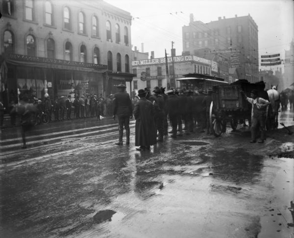 Soldiers from the second Wisconsin regiment line a city street upon returning from Puerto Rico. There is a horse-drawn cart and an electric street car on tracks, as well as a sign on a building that says: "Fred Miller Brewing Co."