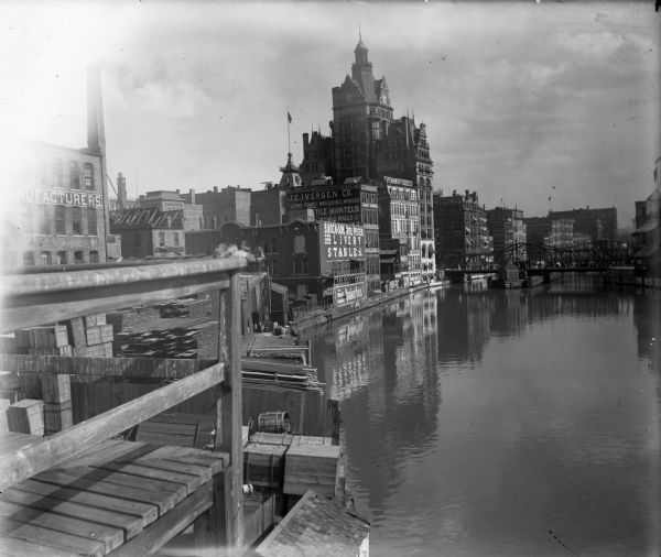 View from pier or dock of buildings along the Milwaukee River, including a building with a clock tower. Signage on brick exteriors reads "J.C. Iverson Co. / Picture Frames, Mouldings, Mirrors," "B. Mock & Son Hotel Pfister Livery Stables," "The Worlds Breakfast, Quaker Oats," and "Espenhain Dry Goods Co. Wholesale & Retail, Carpets & Draperies."