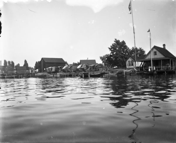 View from water of boats docked near houses on Jones Island. To the right two flags are strung up flagpoles near a building with a covered porch, perhaps a boathouse. Along the shoreline are nets neatly wound up on net reels to dry.