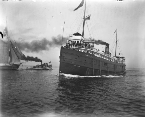 A large steamship, <i>City of Racine</i>, loaded with passengers sails towards Milwaukee Harbor. On the left a tugboat pulls a smaller sailboat out towards Lake Michigan.