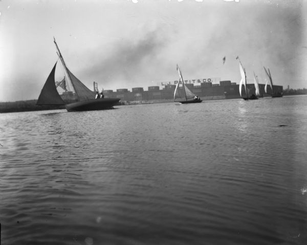 View across water of sailboats in Milwaukee Bay. One of the boats, <i>The Kim</i>, has Syl on board. In the background are industrial buildings, one of which has signage on its roof reading "L.J. Petit & Co."