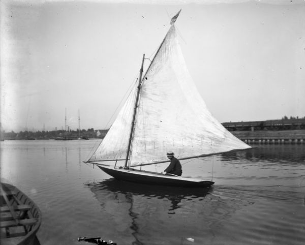 Pennyfeather sitting in a small sailboat in Milwaukee Harbor. There are industrial buildings on the far shoreline, and other boats at anchor in the background.